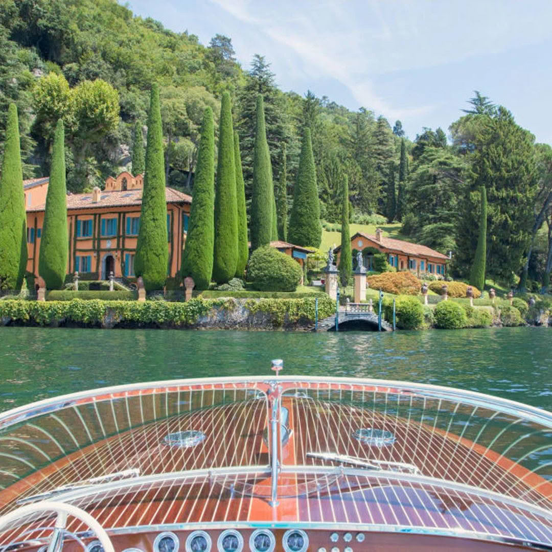 Wooden boat approaching lush lakeside villa with trees