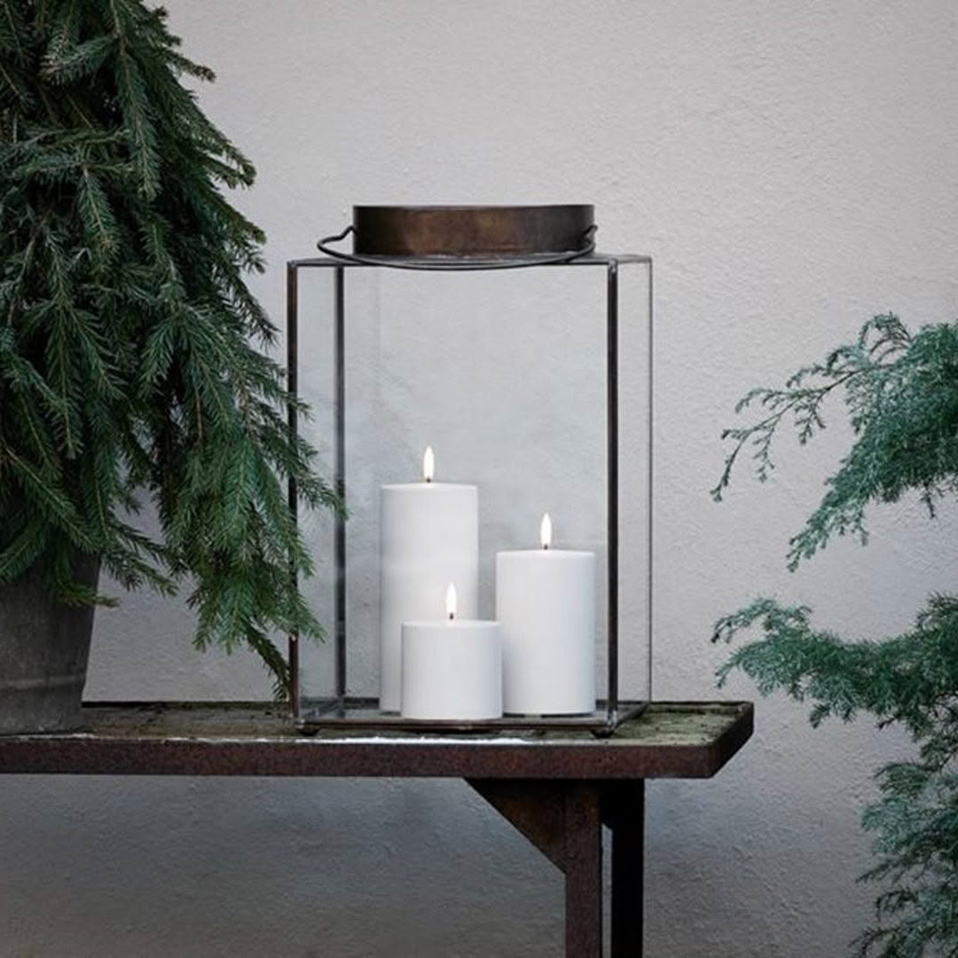 Lit candles in glass lantern on wooden shelf with tree.